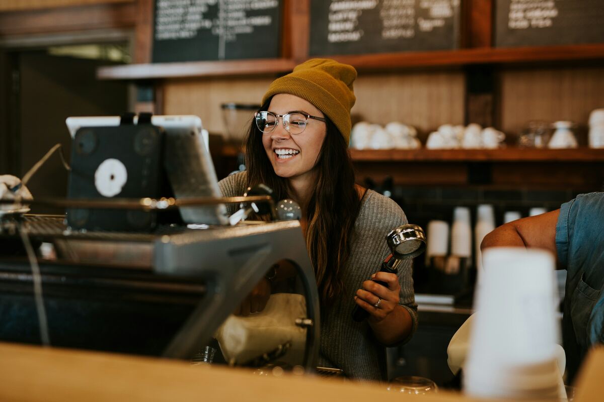 A smiling barista in a mustard beanie holding a portafilter behind a coffee shop counter