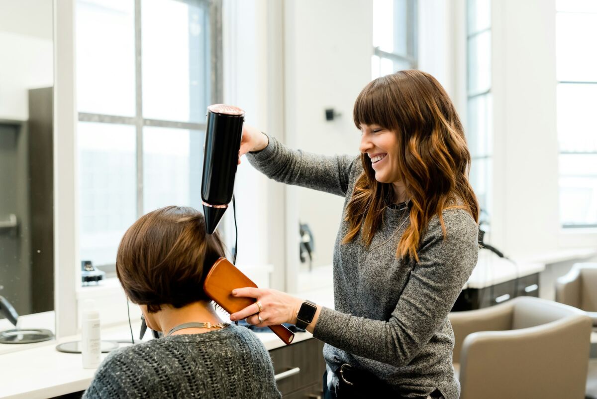 A smiling hairdresser blow-drying a client's hair in a bright salon