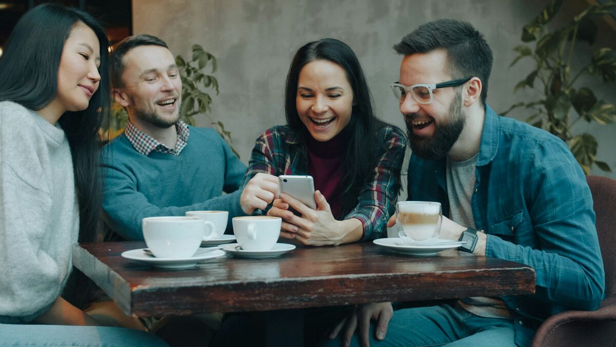Four friends laughing together over a smartphone at a cafe table with coffees